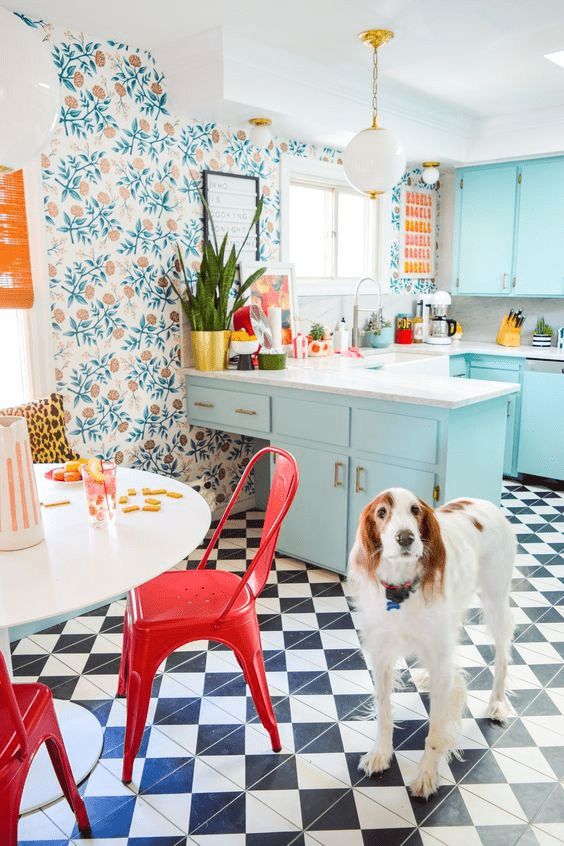 Colorful maximalist kitchen with botanical wallpaper, black-and-white checkered tile flooring, powder blue cabinets, and white or grey countertops. A dog is in the kitchen, staring at the viewer.