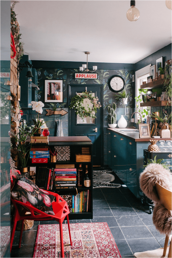 Eclectic maximalist kitchen with bookshelves, plants, teal door frame and cabinets, rugs, and more.